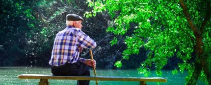 Fotografía de un hombre de avanzada edad, con bastón y boina, sentado en un banco frente a un estanque, en un parque.