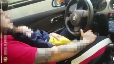 El individuo conduciendo con un bebé en brazos El individuo conduciendo con un bebé en brazos